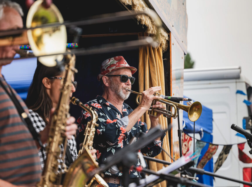 A group of musicians play brass instruments on an outdoor stage. The central figure, wearing sunglasses and a floral shirt, plays the trumpet.