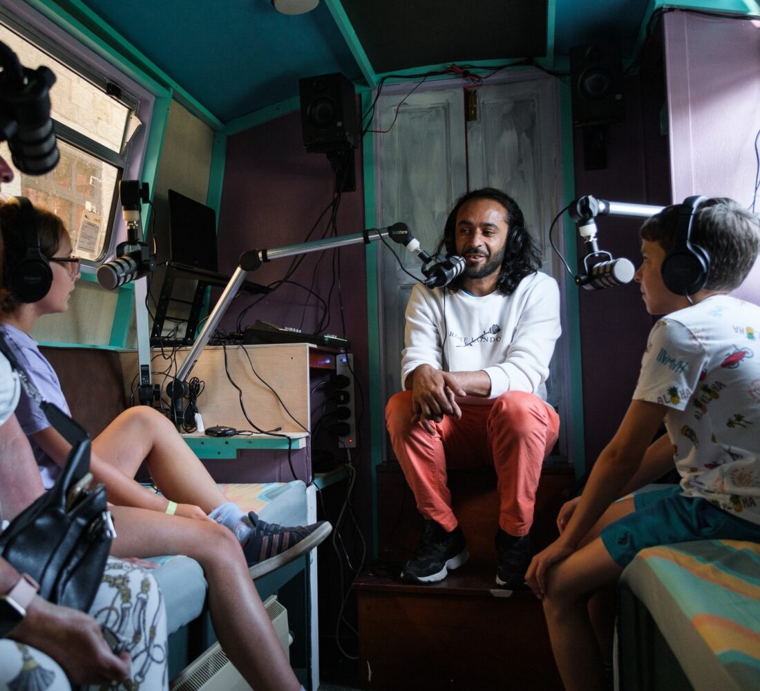 Three people with headphones and microphones sit in a small, colourful studio, having a conversation. The man in the centre speaks while two children sit on either side, listening attentively.