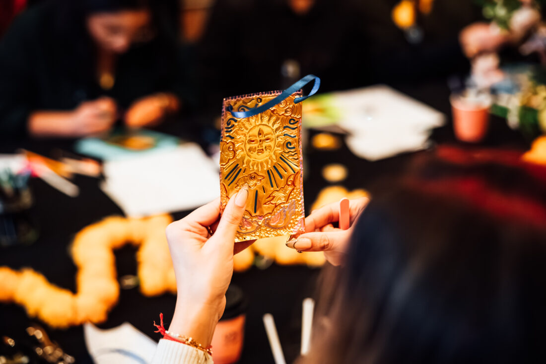 A person holds an embossed gold card with a sun design whilst others work at a table decorated with orange garlands and craft supplies.