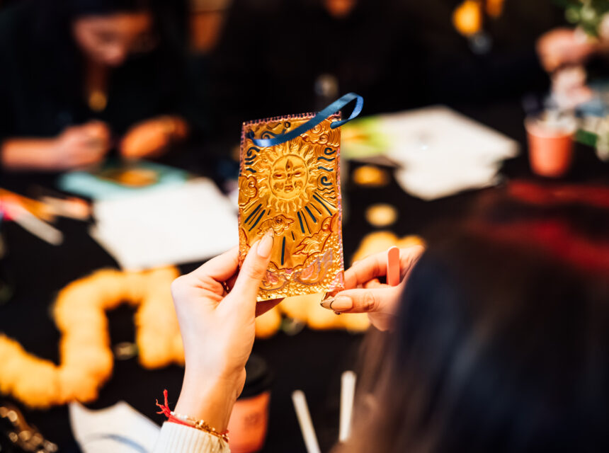 A person holds an embossed gold card with a sun design whilst others work at a table decorated with orange garlands and craft supplies.