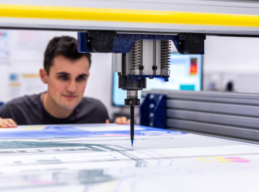 A person closely observes a CNC machine as it engraves or cuts a colourful design on a flat surface in a well-lit workshop.