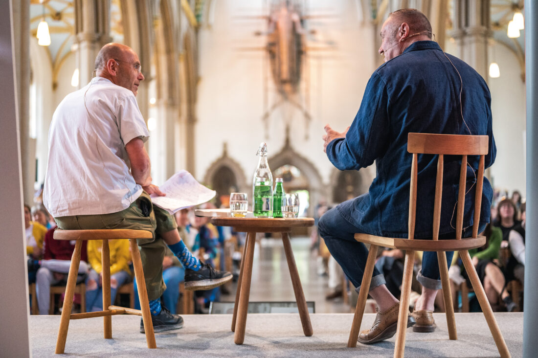 Two people sit on wooden chairs on a stage, facing each other in conversation, with an audience seated in a church-like hall. A table with water bottles and glasses is between them. The background features tall arches.