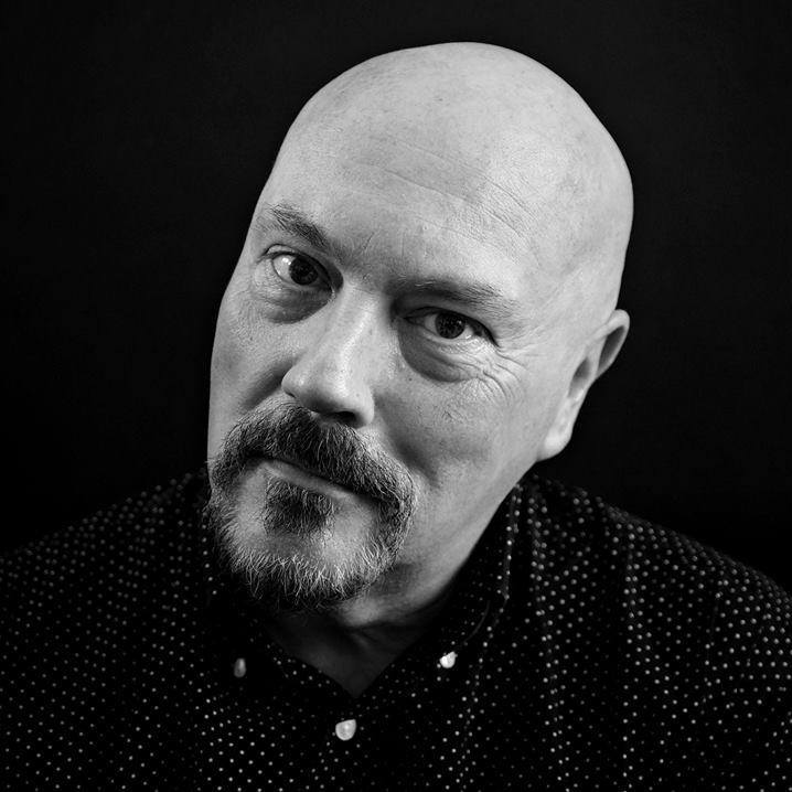 A bald man with a goatee and moustache, wearing a dark, spotted shirt, looks directly at the camera against a plain black background.