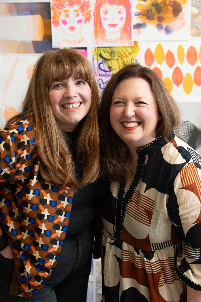 Two women smiling and standing close together in front of a colourful wall filled with vibrant artwork and patterns. Both wear patterned tops and have long hair.