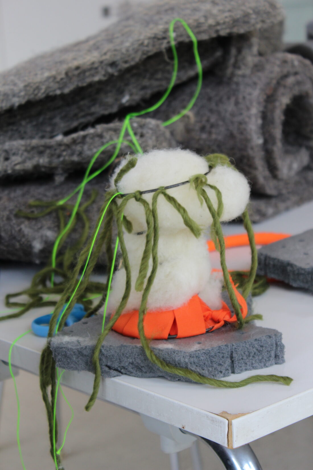 A craft project made of white and orange felt, grey foam, and green yarn sits on a table with grey fabric pieces, blue scissors, and other supplies in the background.