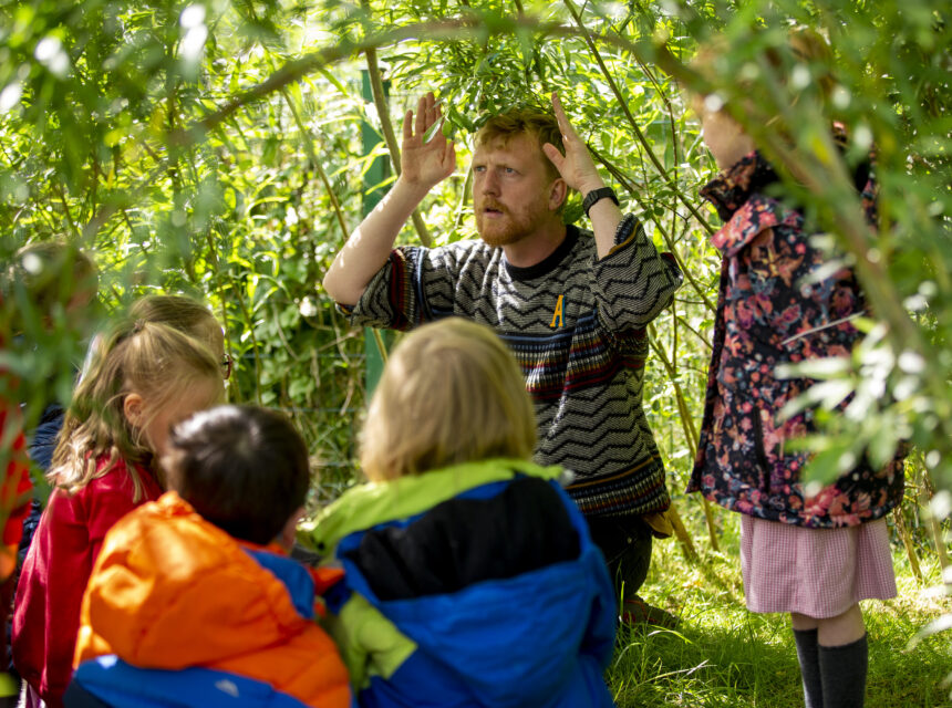 A man sits with a group of children outdoors under green branches, gesturing with his hands above his head, whilst the children listen and watch attentively.