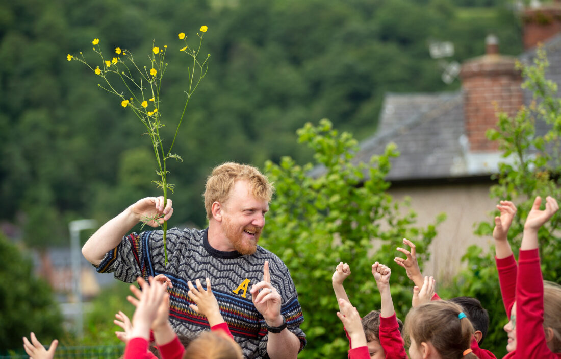 A smiling man holding a tall yellow wildflower stands among children in red uniforms who are reaching up excitedly outdoors, with greenery and a house in the background.