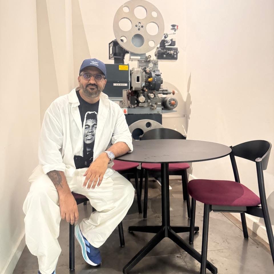 A man in white clothes and a blue cap sits at a round table with red chairs. A vintage film projector is behind him in a minimalist room with light-coloured walls.
