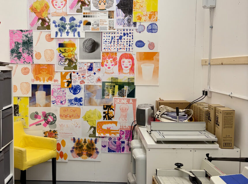 A brightly lit studio space featuring a wall covered in various colourful art prints and drawings. A vibrant yellow armchair sits in the corner, and a desk with a paper cutter and office supplies occupies the foreground.