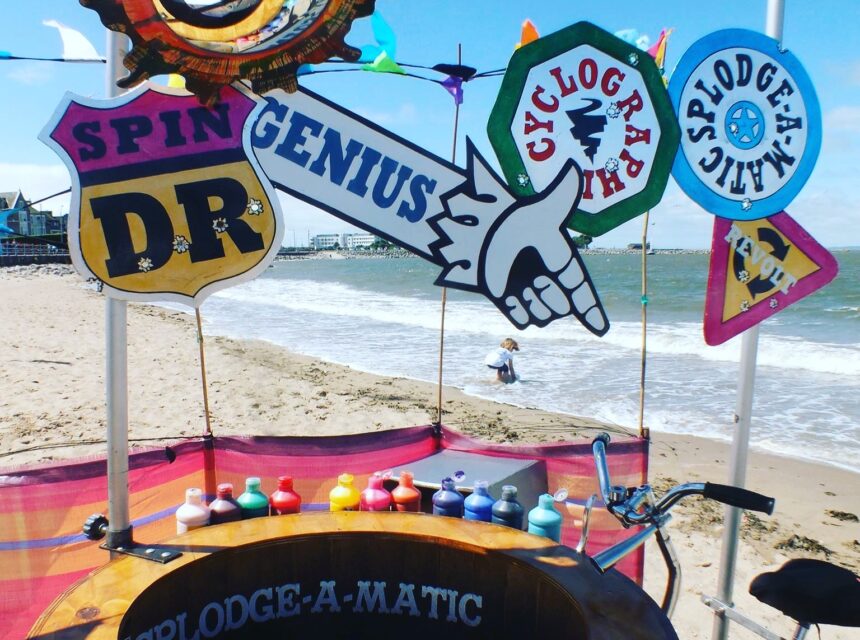 A colourful spin art booth called Splodge-A-Matic is set up on a sandy beach. Paint bottles are arranged in a circle; playful signs say GENIUS and SPINDR. The sea and a child are visible in the background.