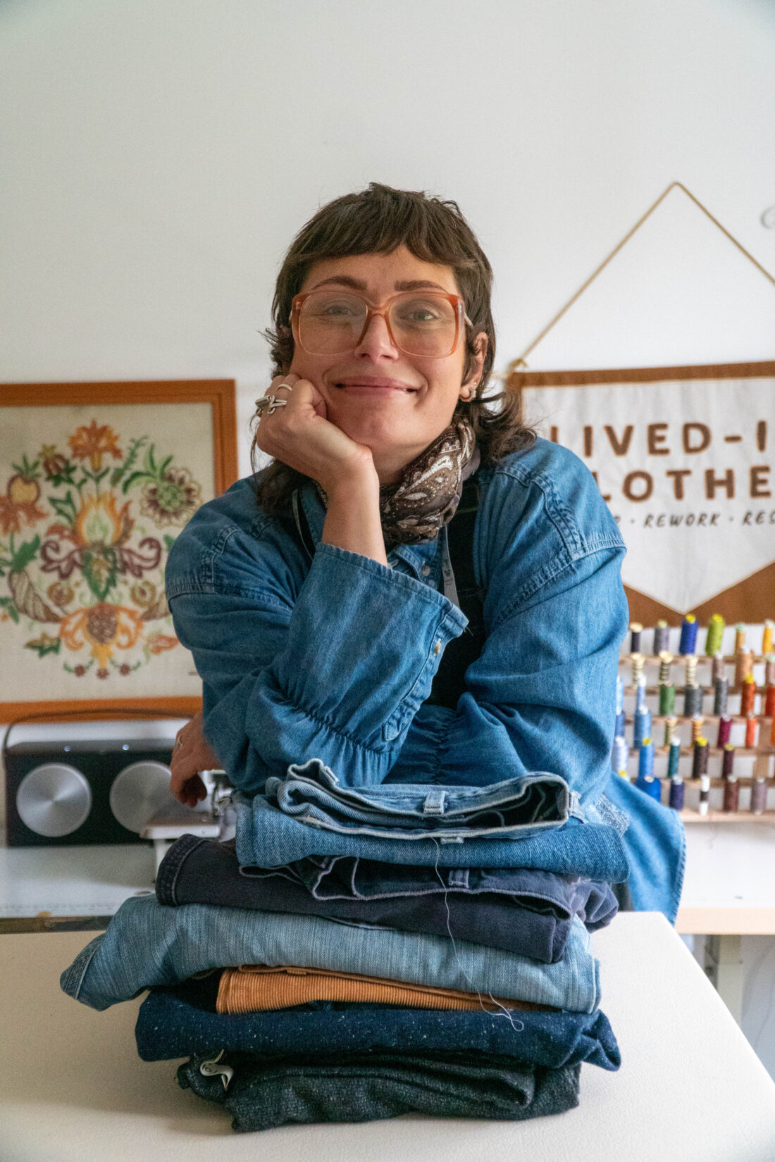 A person in spectacles and denim shirt smiles, resting their chin on their hand above a stack of folded jeans. Sewing supplies and a Lived-In Clothes sign are in the background.