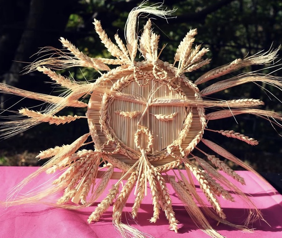 A sun-shaped decoration made from woven straw and wheat sits on a pink surface, with rays and facial features formed by wheat stalks, set against a blurred outdoor background.