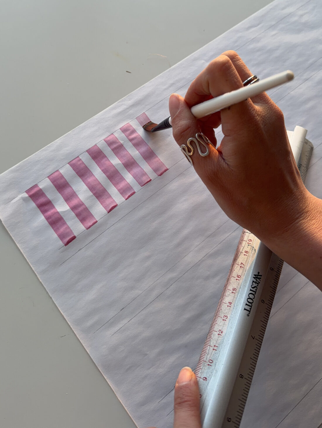 A person paints pink stripes on paper using a brush, with a ruler held in their other hand. The scene is well-lit and the person wears a wavy silver ring.