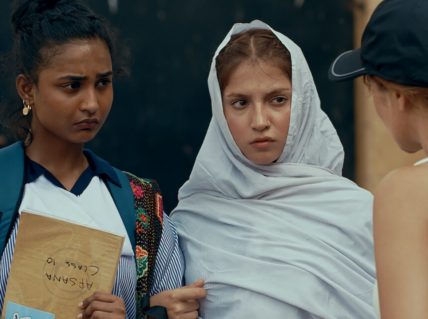 Two young women, one holding a book labelled AB SHAMA CLASS 10, stand closely together looking seriously at someone in a cap who is facing them. One wears a white headscarf, both have intense, concerned expressions.