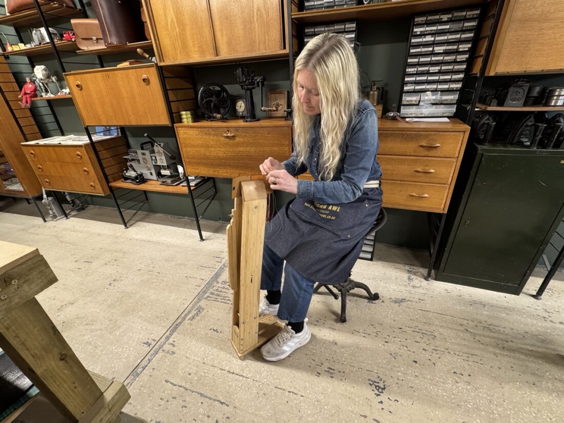 A woman with long blonde hair sits on a stool in a workshop, working on a wooden project. She wears a denim outfit and apron, surrounded by wooden cupboards and various tools.
