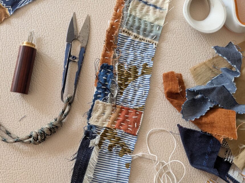 A strip of fabric with patchwork and visible stitches lies on a table surrounded by scissors, thread, fabric offcuts, and sewing tools.