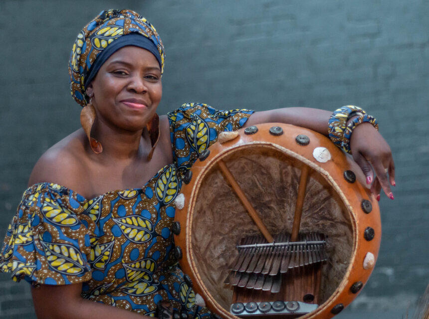 A woman in a vibrant, patterned dress and headwrap smiles whilst holding a large African musical instrument, possibly a kalimba or thumb piano, against a dark green brick wall.
