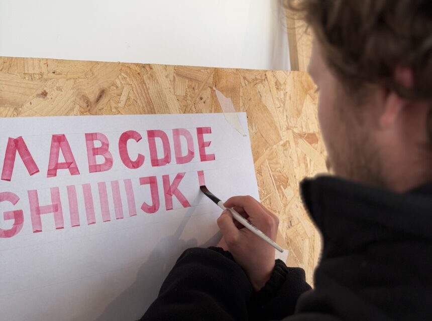 A person in a black jacket paints large pink letters A B C D D E G H I I I I J K on white paper taped to a wooden board. The focus is on the painting hand and letters.