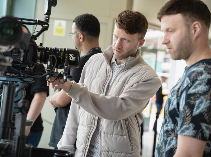 Two men adjust a professional video camera on set, whilst two others stand in the background. One man points at the camera controls, focusing intently.