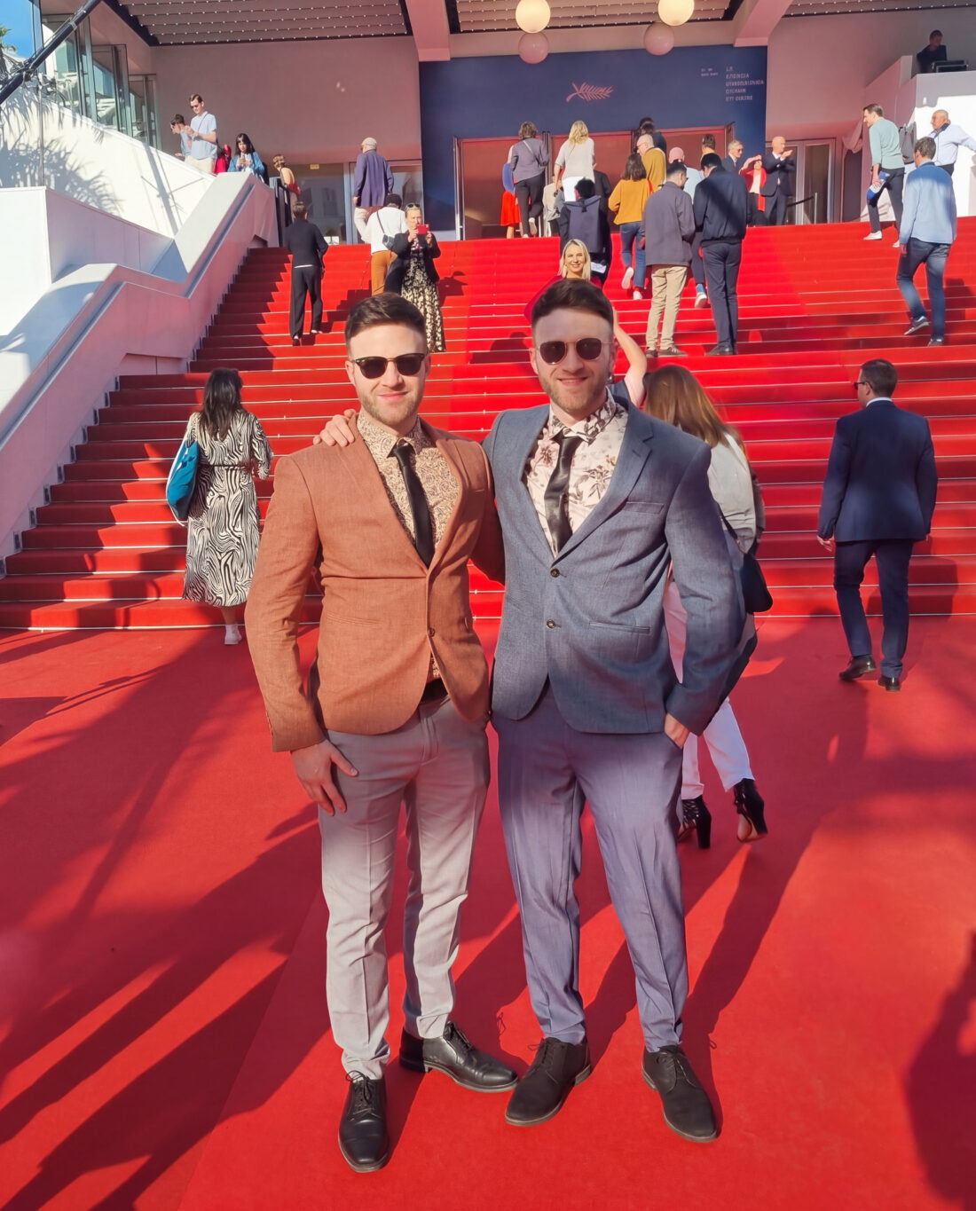 Two men in suits and sunglasses pose together on a sunny red carpet staircase. Other people are walking up and down the steps, and a building entrance is visible in the background.