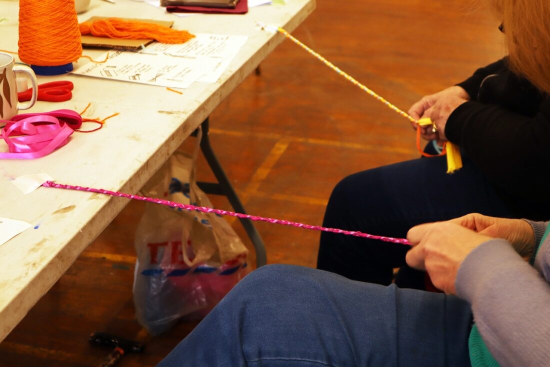 Two people sit at a table, plaiting colourful yarn and ribbon. Supplies, scissors, and printed instructions are on the table. The focus is on their hands and the materials rather than their faces.