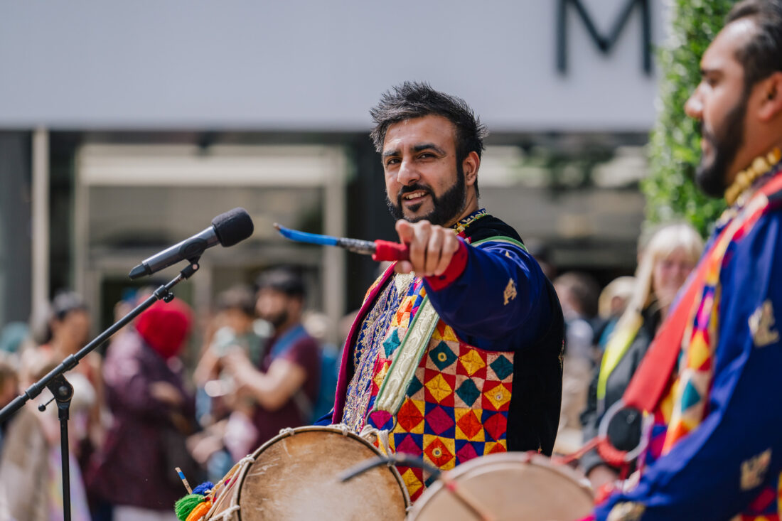 A man in colourful traditional clothing plays a drum and points with a stick, performing outdoors with a microphone nearby. Another drummer stands beside him, and a crowd is gathered in the background.