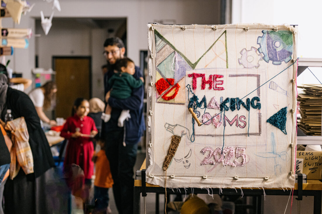Textile artwork reading “The Making Rooms 2025” on a frame, with stitched shapes and drawings; behind, adults and children gather in a busy workshop.