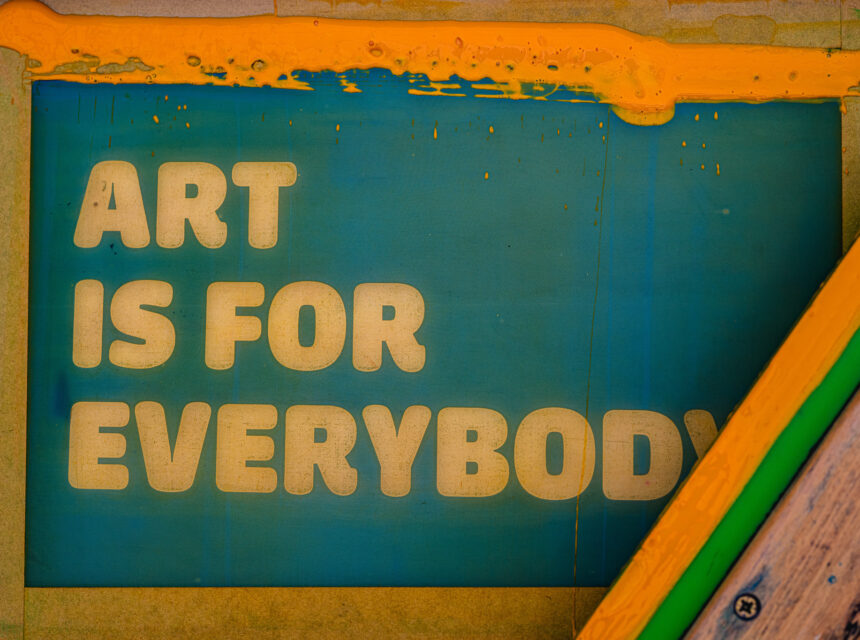 A teal screen displays the yellow words ART IS FOR EVERYBODY, with yellow paint partially dripping over the top and a wooden frame on the lower right side.