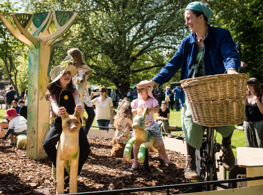 Children ride animal-shaped wooden sculptures at a sunny outdoor fair while an adult pedals a stationary bicycle with a basket, powering the carousel. Trees and people are visible in the background.