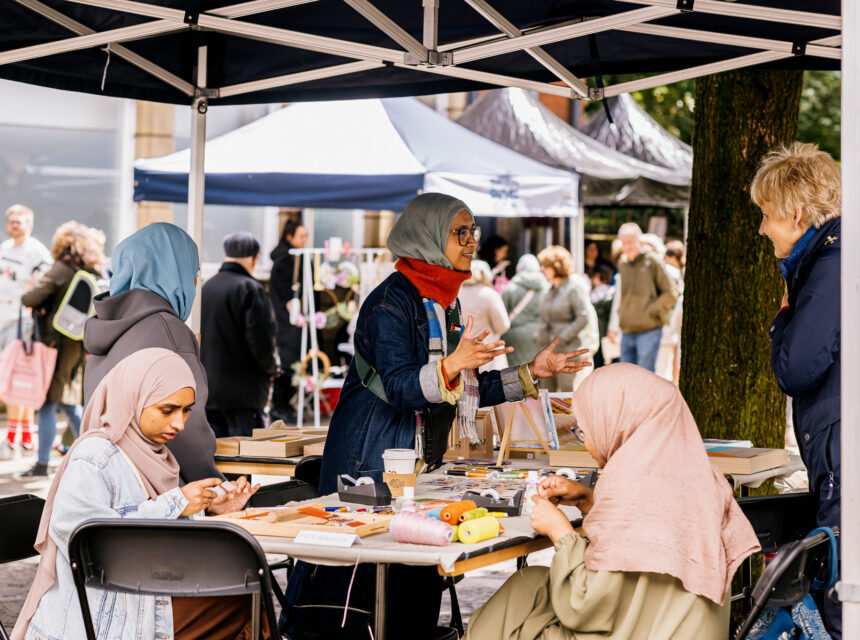 Four women sit and talk at a craft table under a canopy at an outdoor event. Wool, scissors, and craft supplies are on the table. People and tents are visible in the background.