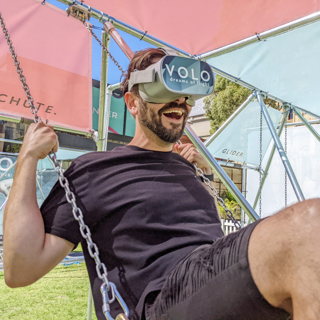 A man in a black T-shirt smiles whilst sitting on a swing, wearing a VR headset labelled VOLO dreams of flight, outdoors under a colourful structure.