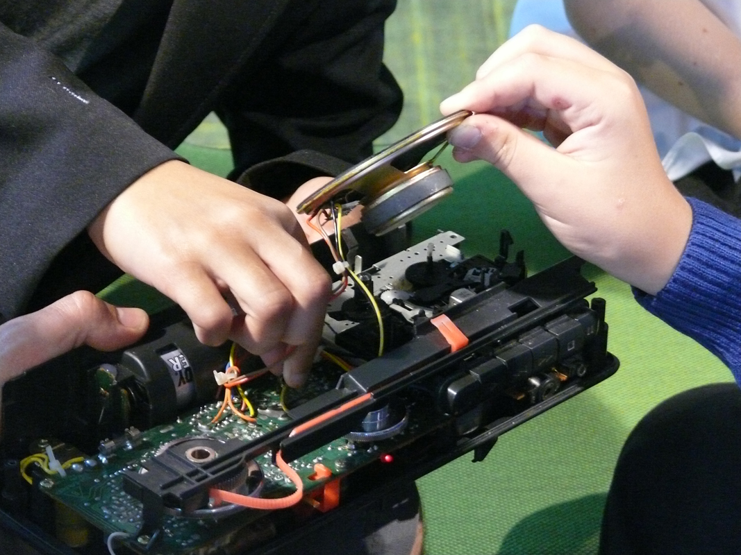 Three hands are working together to repair or assemble an electronic device, possibly a radio or cassette player, with visible wires, circuits, and a loudspeaker. The background is green and slightly blurred.