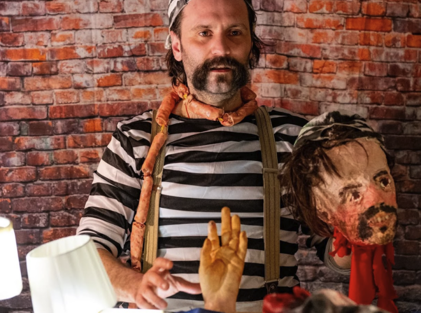 A person in a striped shirt and braces stands in front of a brick wall, holding a realistic puppet head and a prop hand, with lamps illuminating the scene.