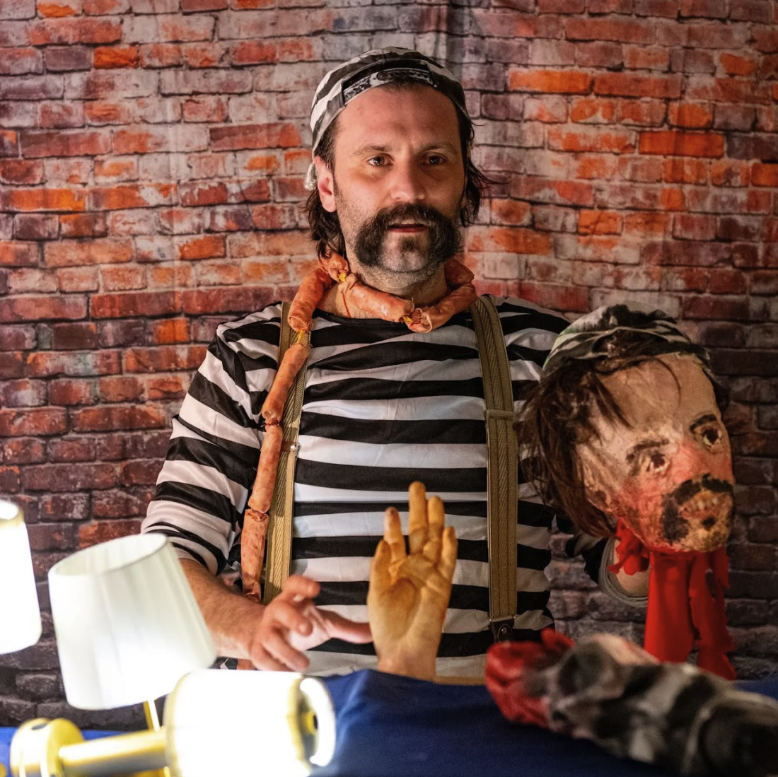 A person in a striped shirt and braces stands in front of a brick wall, holding a realistic puppet head and a prop hand, with lamps illuminating the scene.
