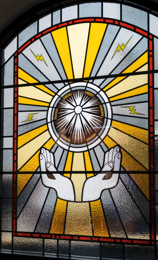 Stained glass window showing two hands uplifted beneath a radiant, circular burst of light with yellow rays and small lightning bolts, symbolising divine or spiritual energy.