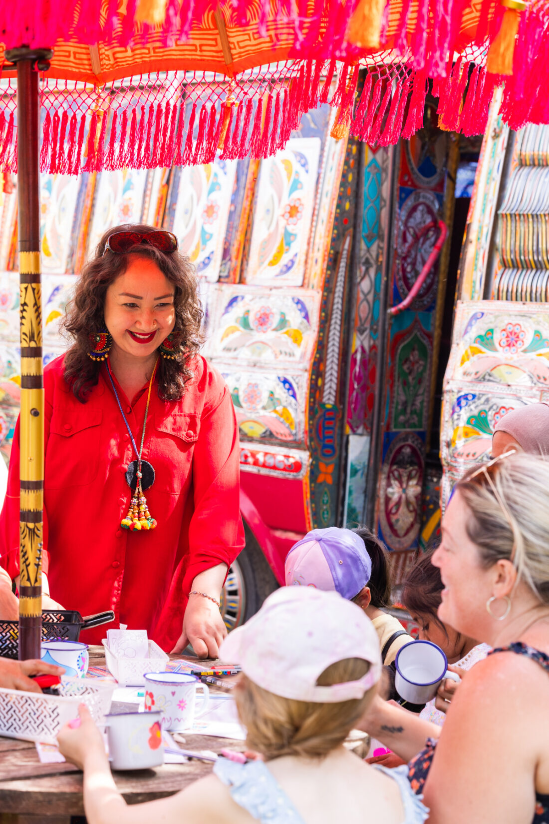 A woman in a red dress smiles whilst leading a crafts activity outdoors with children and adults at a colourful, decorated table under a fringed parasol.