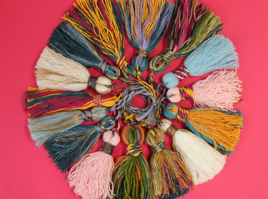 A circular arrangement of colourful yarn tassels on a bright pink background, featuring various textures and colours including blue, yellow, pink, green, and cream.