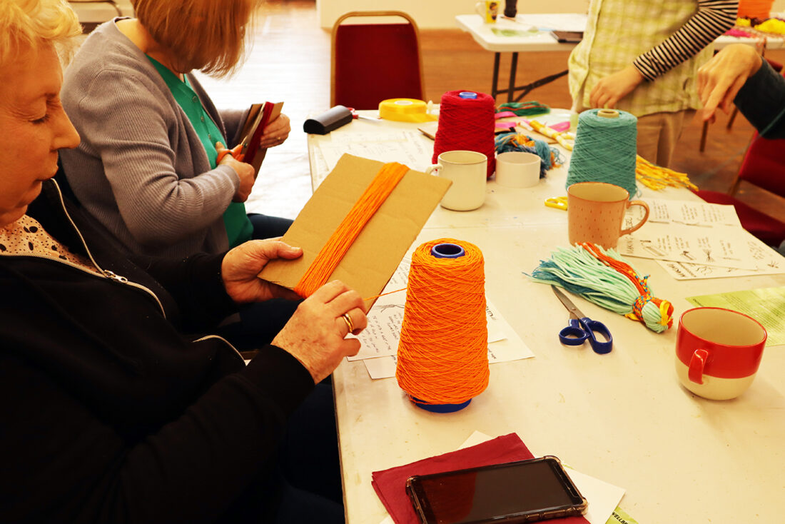 People sit at a table wrapping orange wool around cardboard. Wool cones, scissors, mugs, and papers are on the table. One person holds cardboard with orange wool; another wraps wool. A mobile lies face down in the foreground.