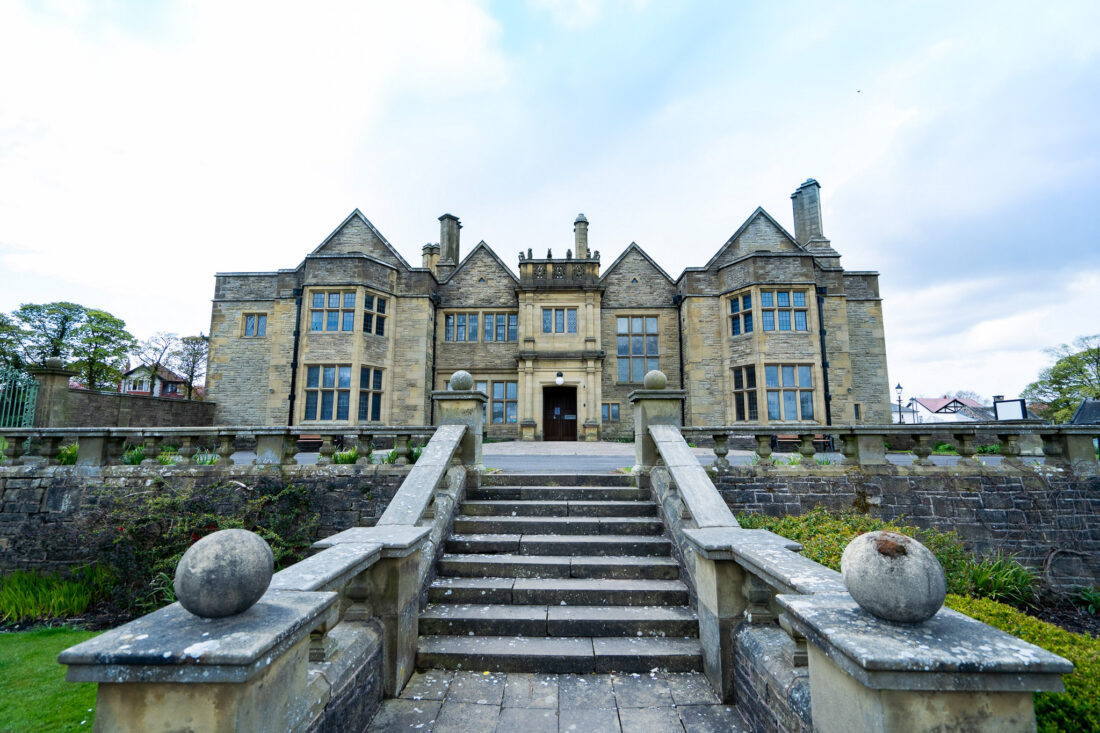 A large, historic stone mansion with multiple chimneys, tall windows, and a grand staircase leading up to the entrance, surrounded by a stone balustrade and landscaped grounds.