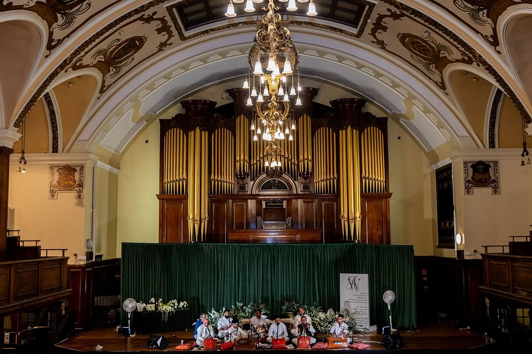 A music group performs on stage in an ornate hall with a high arched ceiling, chandeliers, and a large pipe organ in the background. The stage is decorated with flowers and a green curtain.