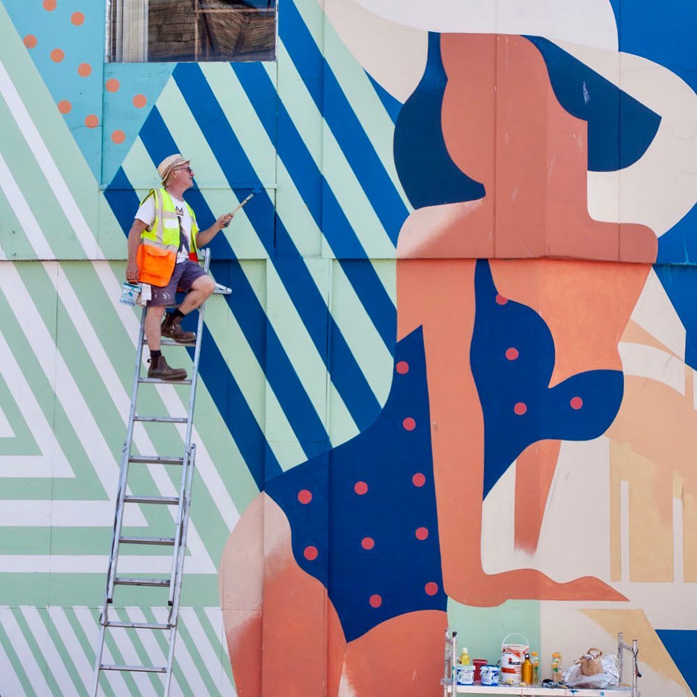 A person on a ladder paints a colourful mural of a woman in a polka dot swimming costume and hat on a wall. Painting supplies are on the ground nearby.