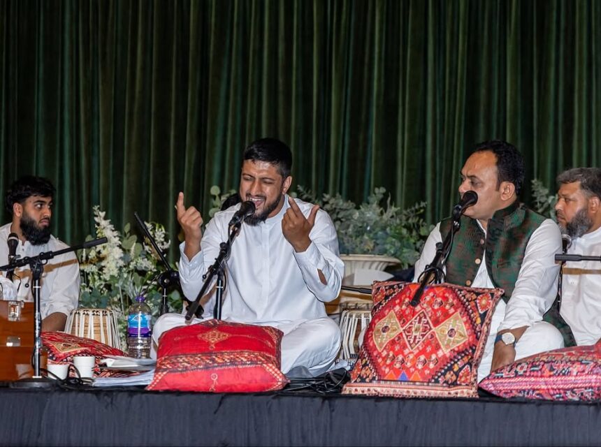 Four men in white traditional attire perform on stage with microphones, cushions, and musical instruments. One man sings passionately, gesturing with his hands, whilst the others attentively accompany him. Green curtains and floral decor are in the background.