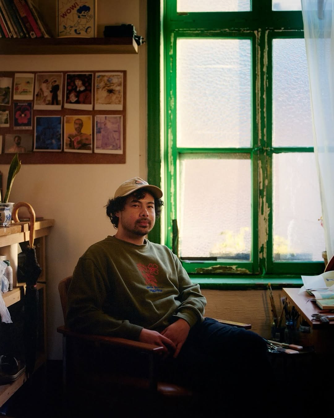 A person wearing a green jumper and cap sits by a desk in a cosy room with art on the wall and a large green-trimmed window letting in soft natural light.