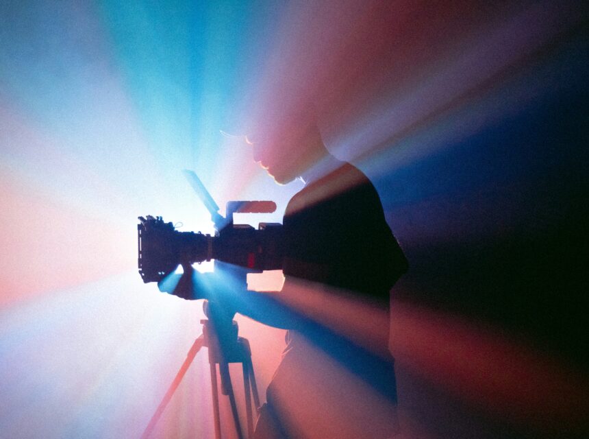 A silhouette of a person operating a camera on a tripod is seen against a dramatic background of colourful, radiating light beams.