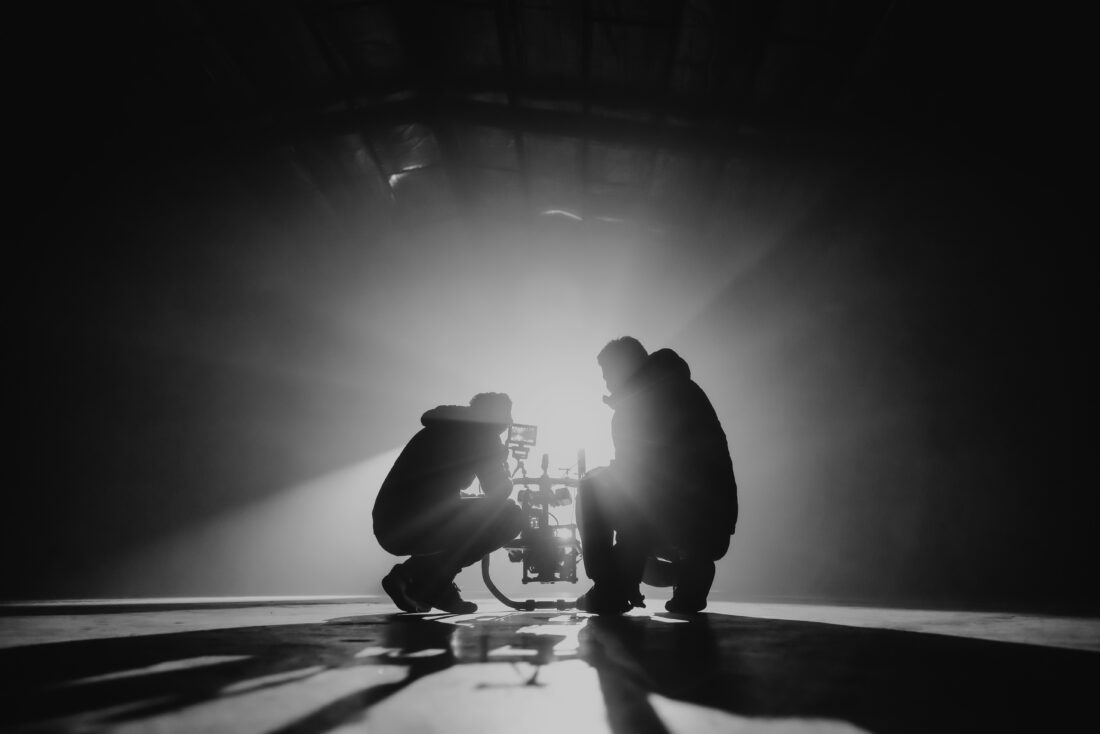 Two people crouch in silhouette around camera equipment in a dark, spacious area with dramatic rays of light shining behind them, creating a striking, high-contrast scene.