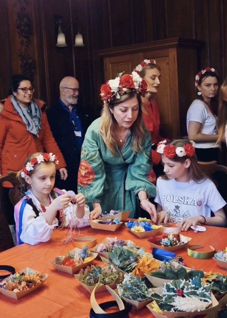 Women and children, many wearing flower crowns, sit and stand around a table making crafts with colourful flowers and greenery in a wood-panelled room. A woman in green helps two girls with their projects.