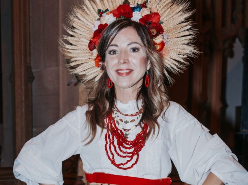 A woman wearing a traditional Eastern European outfit with a white blouse, red skirt, red bead necklaces, and a headdress made of wheat and flowers, smiling and standing indoors.
