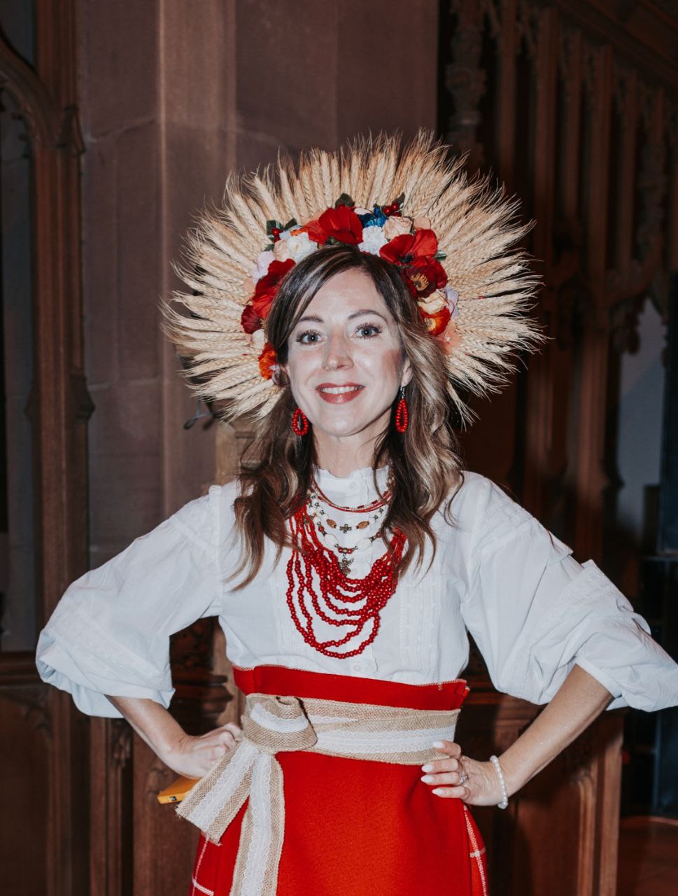 A woman wearing a traditional Eastern European outfit with a white blouse, red skirt, red bead necklaces, and a headdress made of wheat and flowers, smiling and standing indoors.