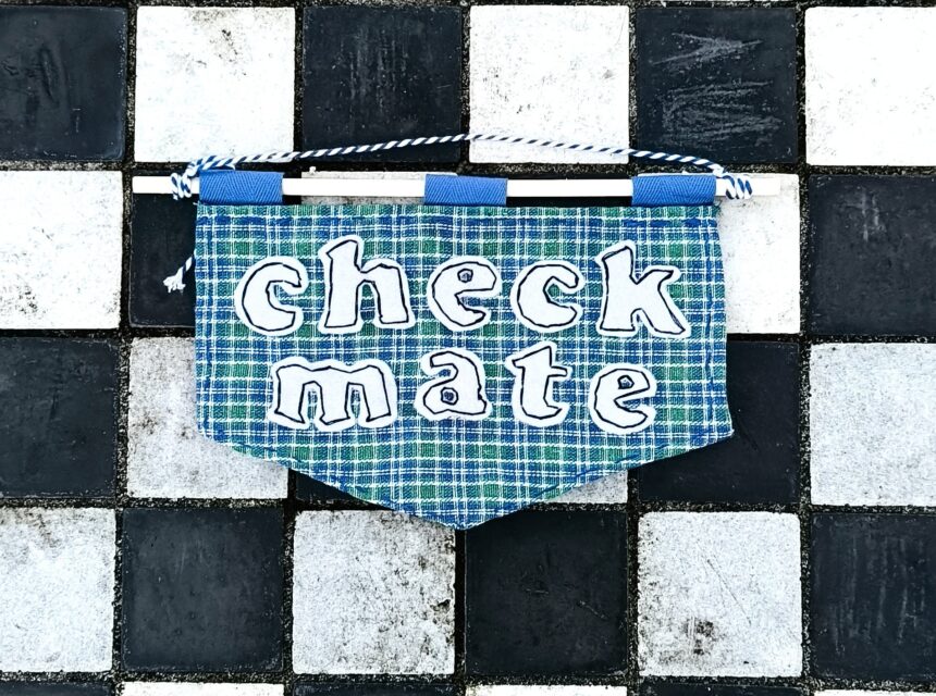 A blue checked banner with the words checkmate hangs in the centre of a black and white chequered tile surface, resembling a chessboard.