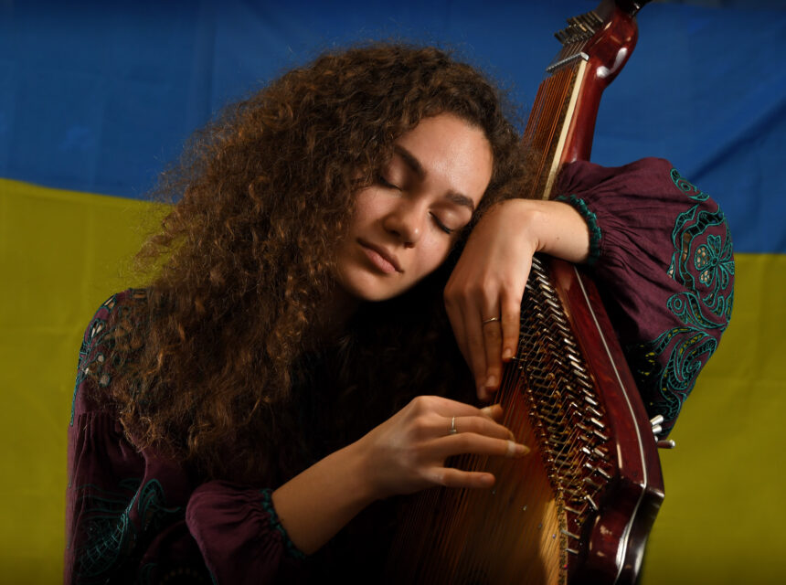 A woman with curly hair plays a harp-like instrument with her eyes closed, wearing a purple embroidered top, in front of a blue and yellow flag.